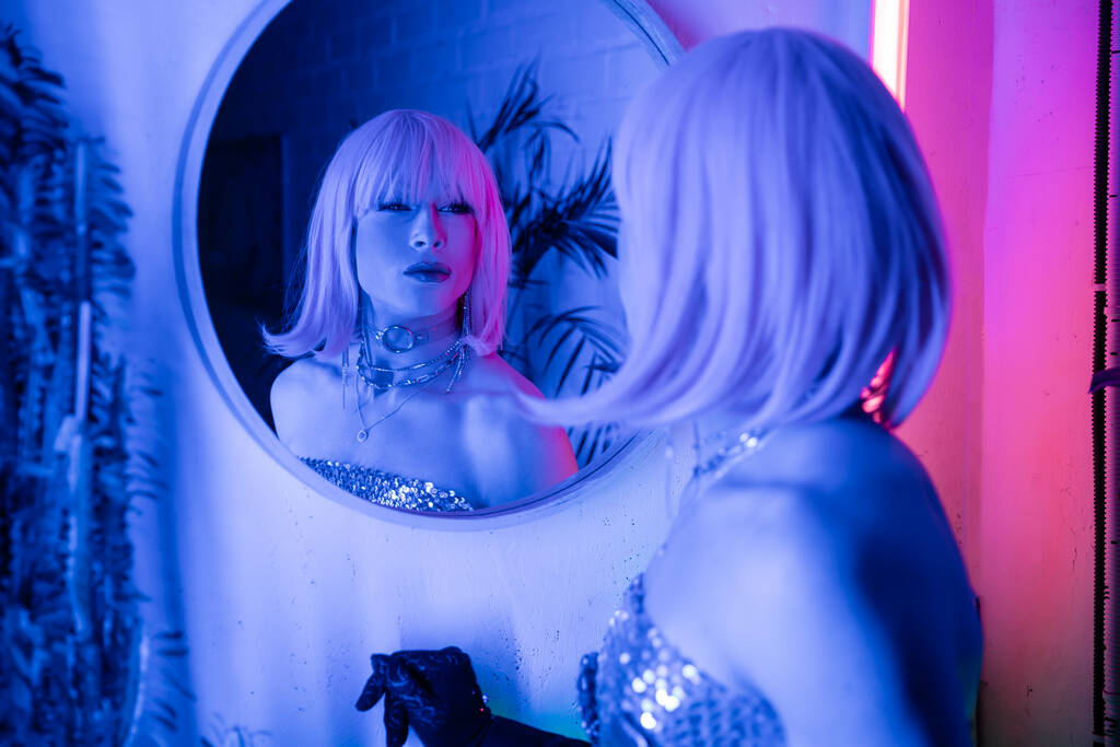 Θολή drag queen σε περούκα και κορυφή κοιτάζοντας τον καθρέφτη σε νέον φως στο σπίτι  - Φωτογραφία, εικόνα