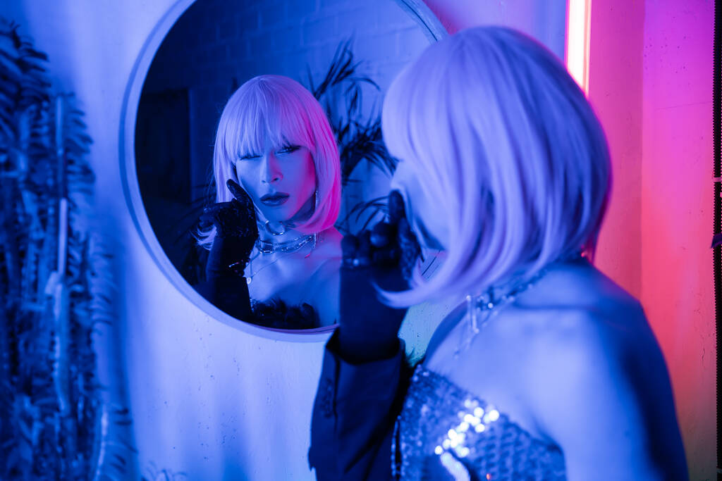 Fashionable drag queen in wig looking at mirror in neon light at home  - Photo, Image