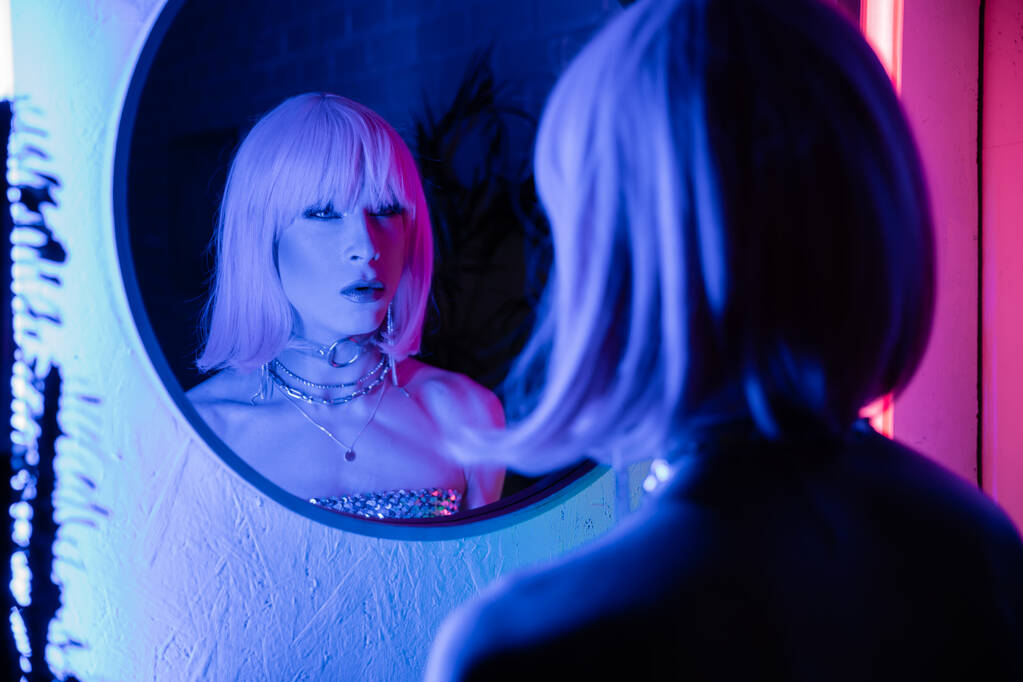 Elegante drag queen in parrucca guardando lo specchio vicino alla luce al neon a casa  - Foto, immagini