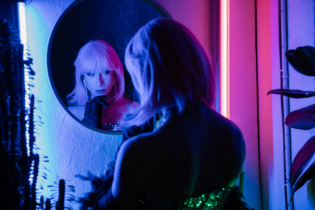 Modieuze drag queen kijken naar spiegel terwijl staan in de buurt van planten en neon licht thuis   - Foto, afbeelding
