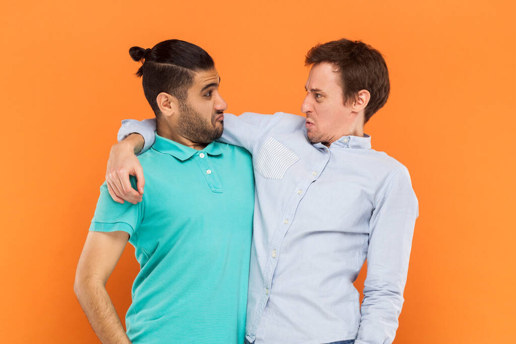 Ritratto di due amici uomini comici in piedi e abbracciati, che si guardano con buffe espressioni facciali accigliate. Indoor studio girato isolato su sfondo arancione. - Foto, immagini