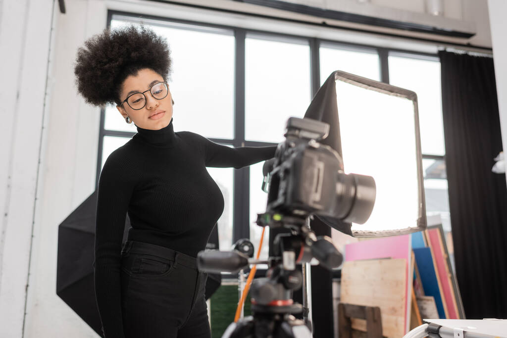 Afrikanisch-amerikanischer Content-Hersteller mit Brille blickt auf verschwommene Digitalkamera, während er in der Nähe von Scheinwerfern im Fotostudio steht - Foto, Bild