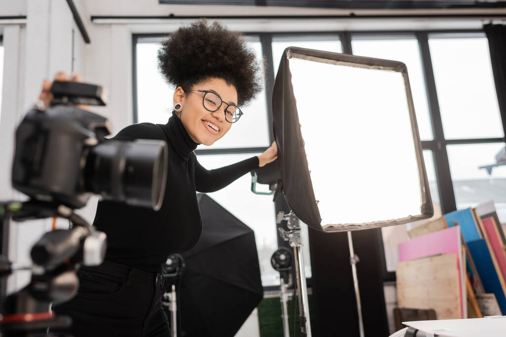 alegre africano americano contenido fabricante sonriendo a cámara cerca reflector y borrosa cámara digital en estudio de fotos - Foto, Imagen