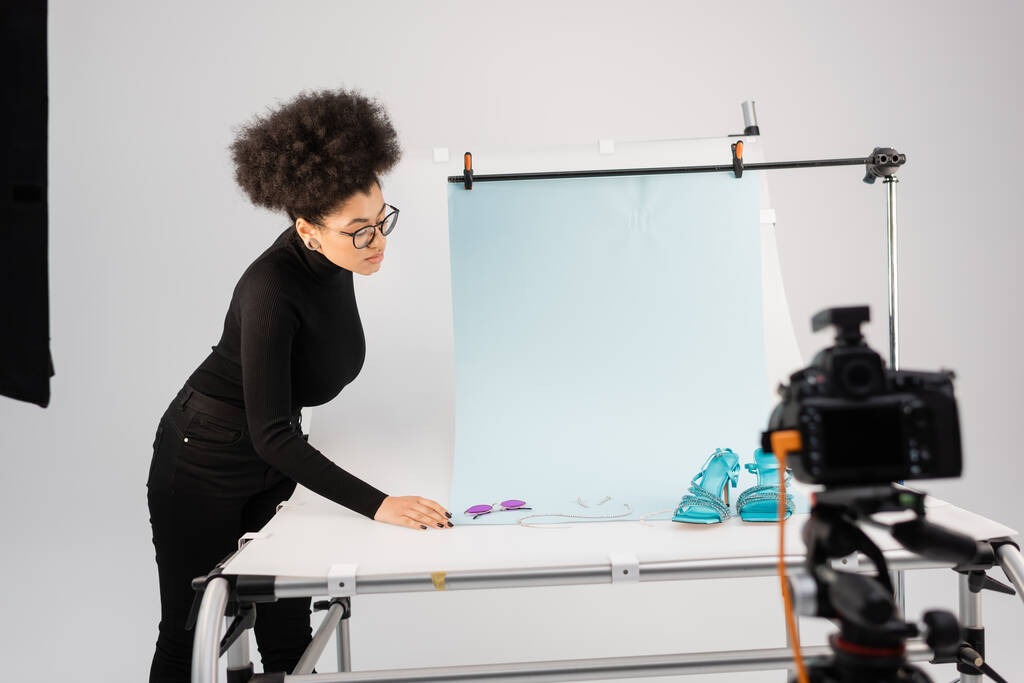 Африканский производитель контента смотрит на стильные солнцезащитные очки и сандалии на столе для съемок рядом с размытой цифровой камерой в фотостудии - Фото, изображение