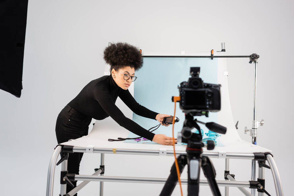 Αφρικανική αμερικανική μηχανή περιεχομένου κοιτάζοντας θολή ψηφιακή φωτογραφική μηχανή, ενώ κρατώντας μετρητή έκθεσης κοντά γυρίσματα τραπέζι στο σύγχρονο στούντιο φωτογραφιών - Φωτογραφία, εικόνα