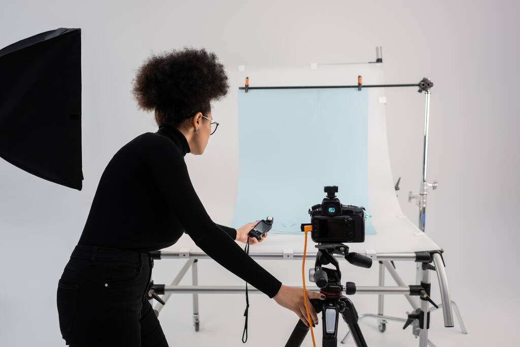 Αφροαμερικανός παραγωγός περιεχομένου με μετρητή έκθεσης κοντά σε ψηφιακή φωτογραφική μηχανή και φωτογράφηση σε σύγχρονο στούντιο - Φωτογραφία, εικόνα