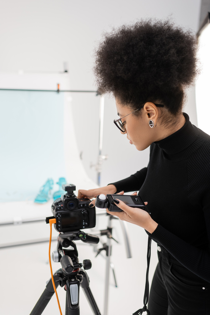vue latérale du producteur de contenu afro-américain avec compteur d'exposition ajustant l'appareil photo numérique moderne dans le studio photo - Photo, image