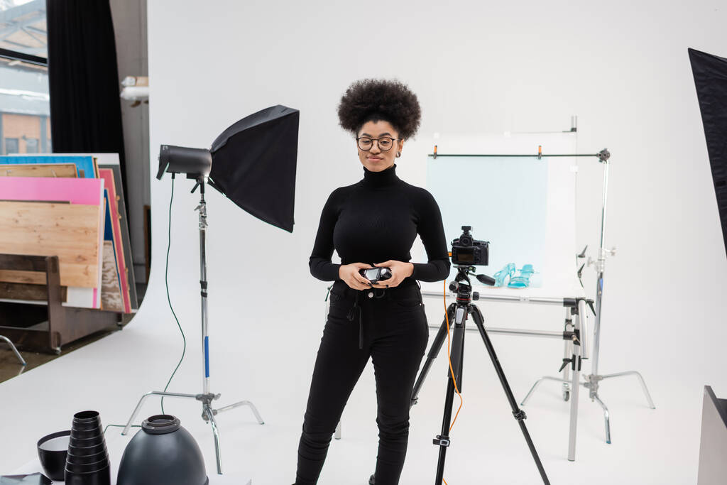 uśmiechnięty afrykański producent treści stojący z miernikiem ekspozycji w pobliżu aparatu cyfrowego i sprzętu oświetleniowego w nowoczesnym studiu fotograficznym - Zdjęcie, obraz