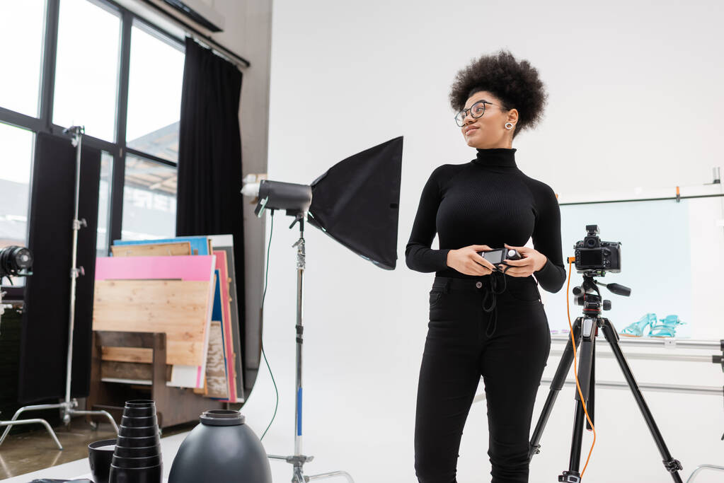 africký americký producent obsahu s měřičem expozice při pohledu mimo softbox reflektor a digitální fotoaparát ve fotostudiu - Fotografie, Obrázek
