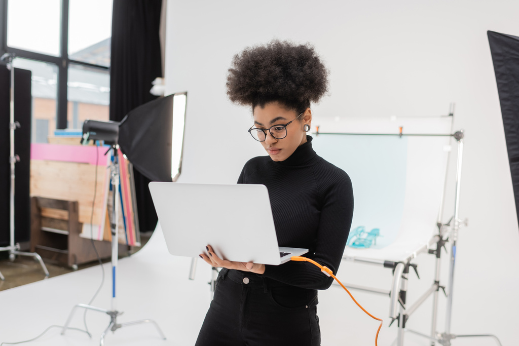 Amerykański producent treści afrykańskich w okularach oglądający laptopa w pobliżu reflektora softbox i stołu strzeleckiego w studiu fotograficznym - Zdjęcie, obraz