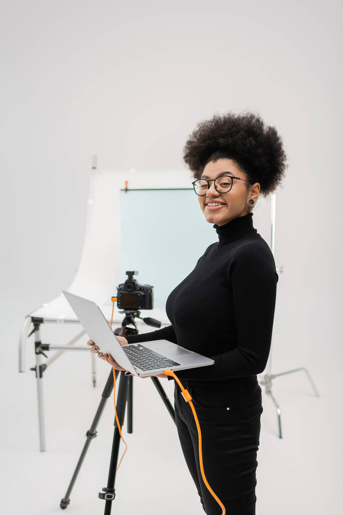 Gözlüklü, dizüstü bilgisayarlı ve fotoğraf stüdyosundaki kameraya bakan mutlu Afro-Amerikan yapımcı. - Fotoğraf, Görsel