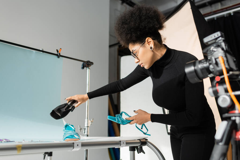 Afro-Amerikan içerikli yapımcı çekim masasının yanında moda ayakkabılar tutuyor ve fotoğraf stüdyosunda bulanık dijital kamera var. - Fotoğraf, Görsel