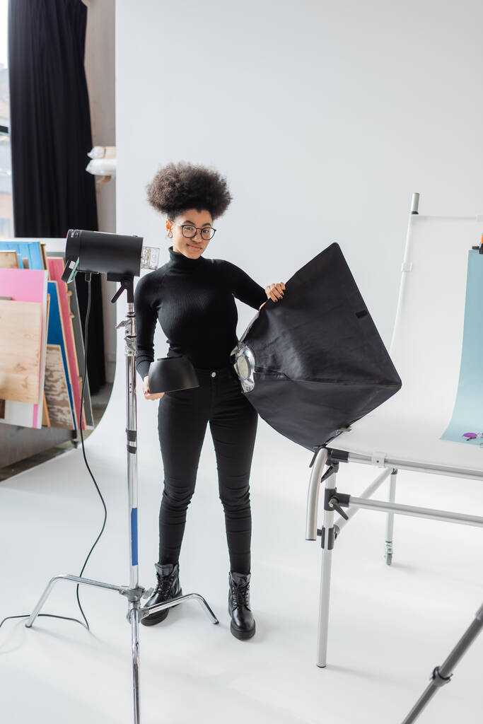 in voller Länge lächelnder afrikanisch-amerikanischer Content Maker in schwarzer Kleidung mit Softbox in der Nähe von Scheinwerfern im Fotostudio - Foto, Bild
