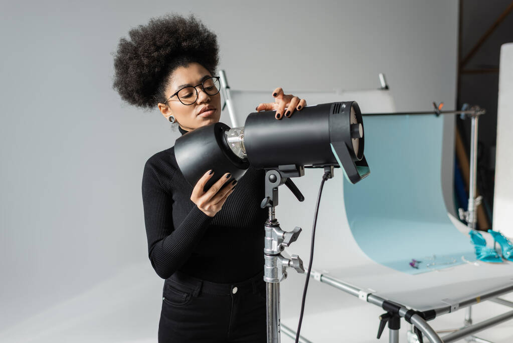 Afrikaans-Amerikaanse content manager in bril en zwarte coltrui werken met stroboscoop lamp in de buurt van fototafel in fotostudio - Foto, afbeelding