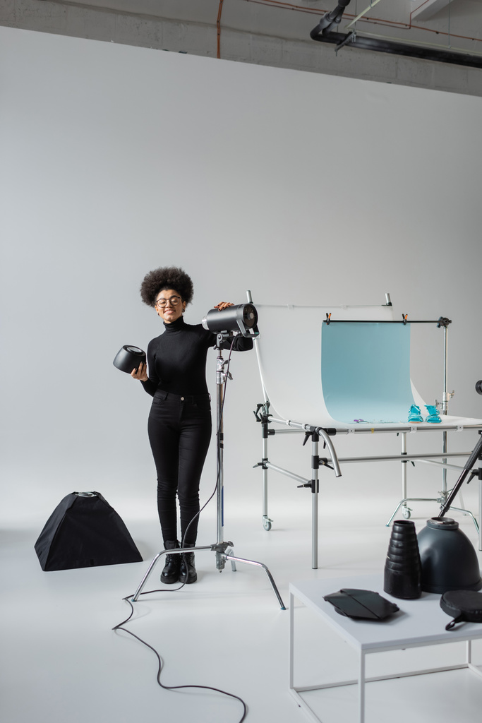 full length of happy african american content maker near strobe lamp and shooting table in contemporary photo studio - Photo, Image