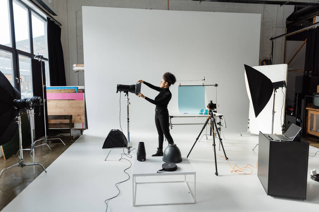 comprimento total do produtor de conteúdo americano africano em roupas pretas montando equipamentos de iluminação em estúdio de fotos espaçoso - Foto, Imagem