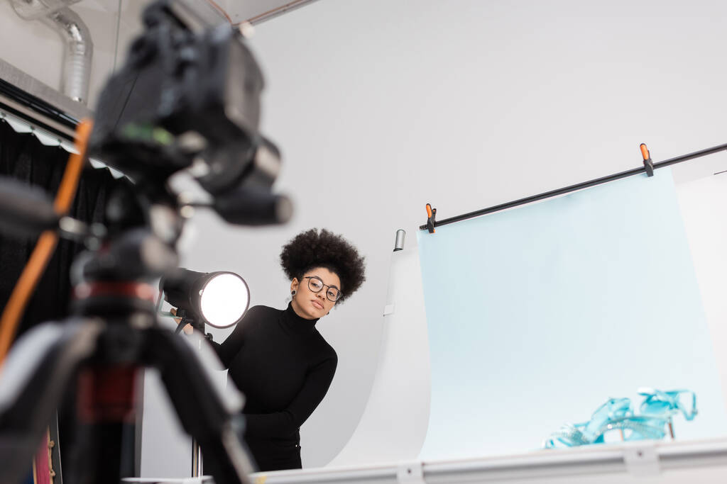 アフリカ系アメリカ人のコンテンツメーカーがスポットライト近くのぼやけたデジタルカメラを見て、写真スタジオで流行の靴でテーブルを撮影 - 写真・画像