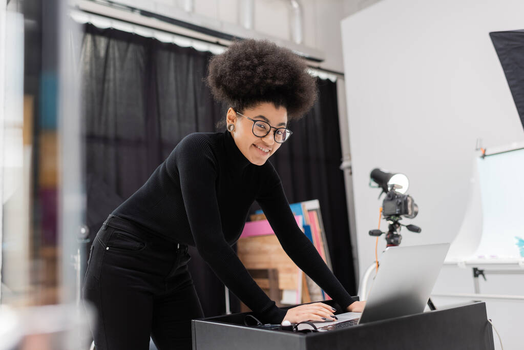 fabricant de contenu afro-américain élégant et joyeux en regardant la caméra et en utilisant un ordinateur portable dans le studio photo - Photo, image