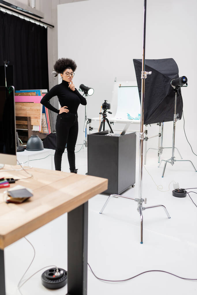 pensive afrikkalainen amerikkalainen sisällöntuottaja seisoo käsi lantiolla lähellä kannettavaa tietokonetta ja softbox heijastin valokuva studio - Valokuva, kuva