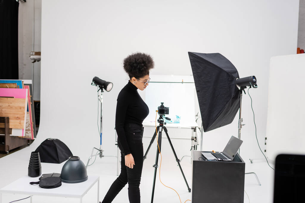 side view afro-amerikai tartalom termelő fekete ruhák közelében laptop és világítás berendezések modern fotó stúdió - Fotó, kép