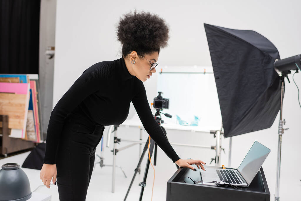 боковой вид африканского производителя контента с помощью ноутбука с Blank-экраном рядом с мыльным боксом в фотоателье - Фото, изображение