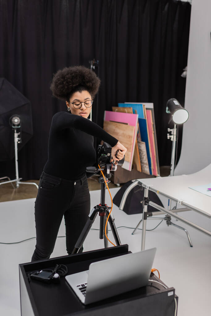 Gözlüklü Afro-Amerikan içerik yapımcısı fotoğraf stüdyosunda dizüstü bilgisayarın yanında dijital kamera monte ediyor. - Fotoğraf, Görsel