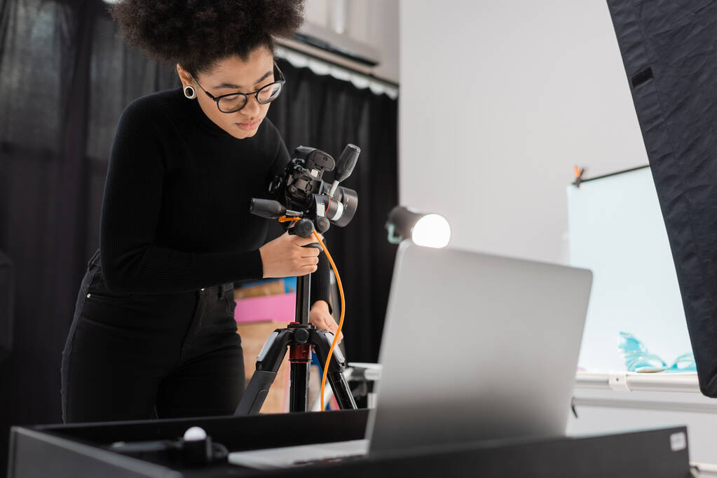producteur de contenu afro-américain ajuster appareil photo numérique sur trépied près d'ordinateur portable flou dans le studio photo - Photo, image
