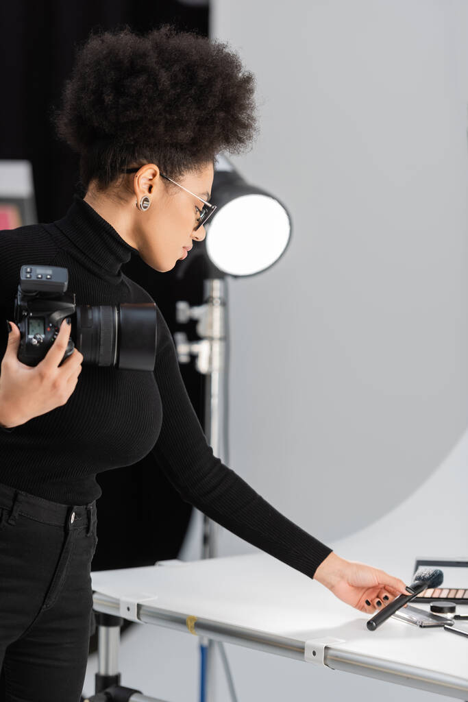 Afro-Amerikan içerik yapımcısı dijital kamerasıyla kozmetik fırçasını fotoğraf stüdyosundaki çekim masasında tutuyor. - Fotoğraf, Görsel