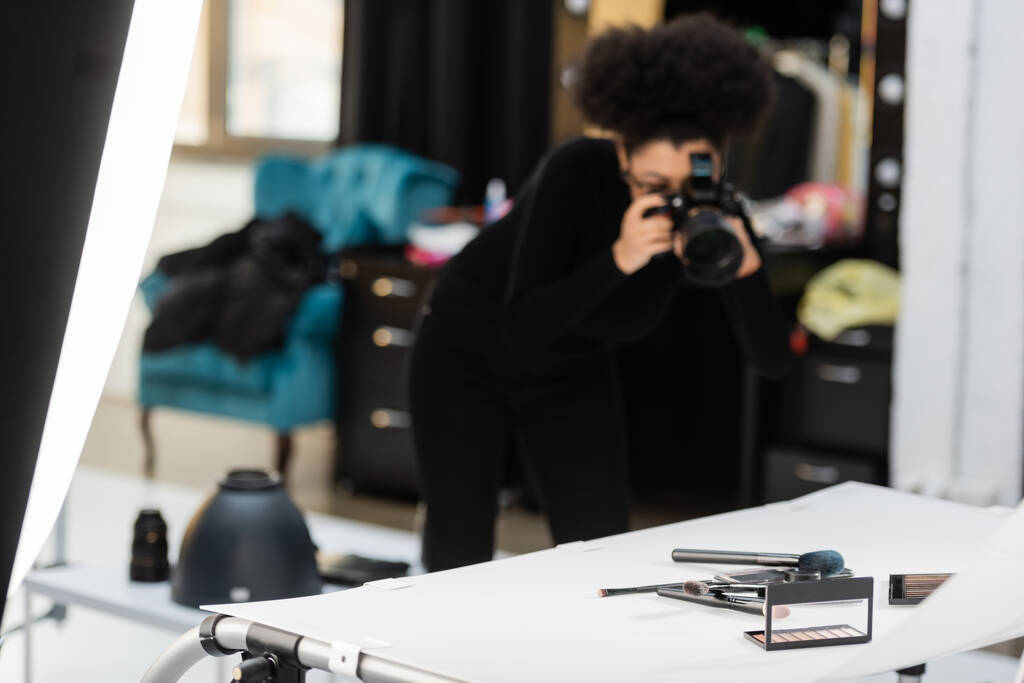 blurred african american content maker taking photo of beauty tools and decorative cosmetics on shooting table in studio - Photo, Image