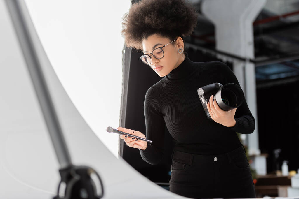 afrikaanse amerikaanse content manager met digitale camera en cosmetische borstel in fotostudio - Foto, afbeelding
