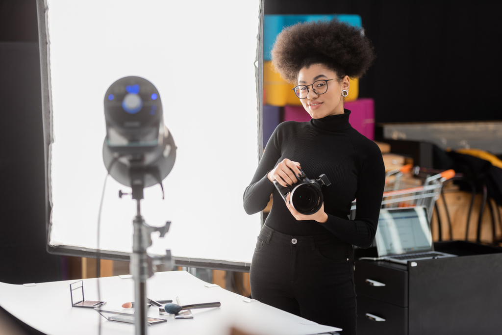 fabricante de contenido afroamericano alegre con cámara digital sonriendo cerca foco borroso en estudio de fotos - Foto, imagen