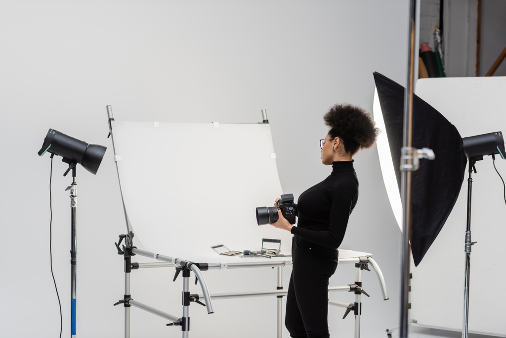 widok z boku afrykański amerykański content maker z aparatem cyfrowym patrząc na dekoracyjne kosmetyki i narzędzia kosmetyczne w studio fotograficznym - Zdjęcie, obraz
