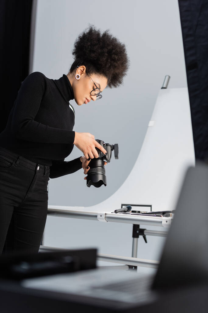 africký americký fotograf v černém roláku a brýle při pohledu na digitální fotoaparát v blízkosti dekorativní kosmetiky ve fotostudiu - Fotografie, Obrázek