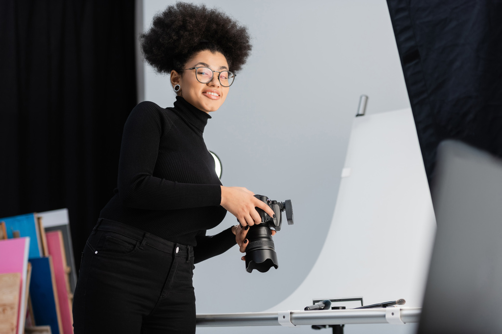 装飾的な化粧品や写真スタジオの美容器具の近くのカメラで笑顔眼鏡で楽しいアフリカ系アメリカ人の写真家 - 写真・画像