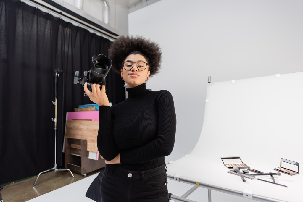 souriant producteur de contenu afro-américain tenant appareil photo numérique tout en se tenant à la table de tir avec des cosmétiques décoratifs en studio photo - Photo, image