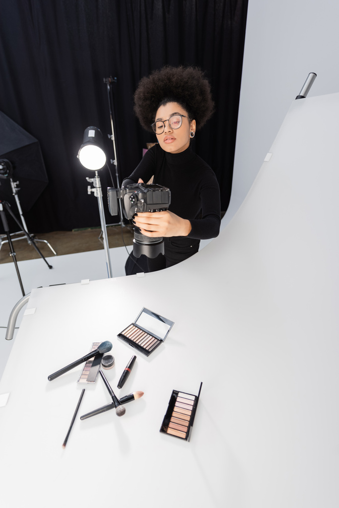 africký americký fotograf v brýlích fotografování kosmetických nástrojů a kosmetiky na fotografickém stole - Fotografie, Obrázek