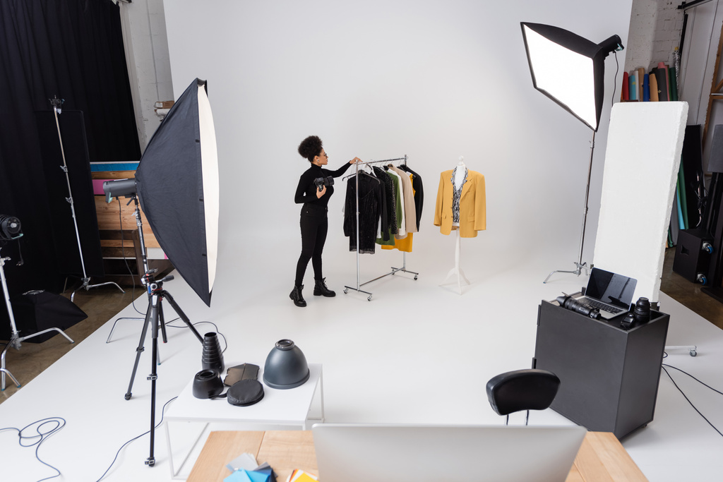 comprimento total do produtor de conteúdo afro-americano trabalhando com nova coleção de roupas elegantes perto de holofotes no estúdio de fotos - Foto, Imagem