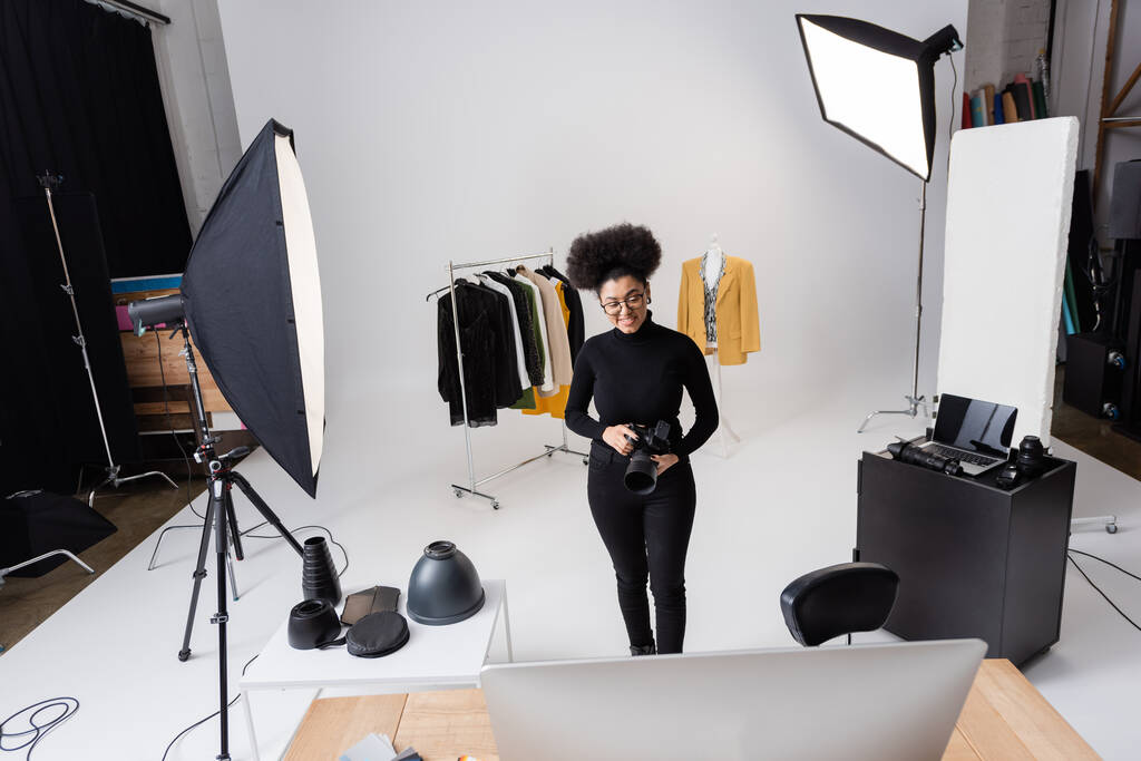 χαρούμενος αφρικανός παραγωγός περιεχομένου με ψηφιακή κάμερα κοντά σε μοντέρνα ρούχα και εξοπλισμό φωτισμού στο φωτογραφικό στούντιο - Φωτογραφία, εικόνα