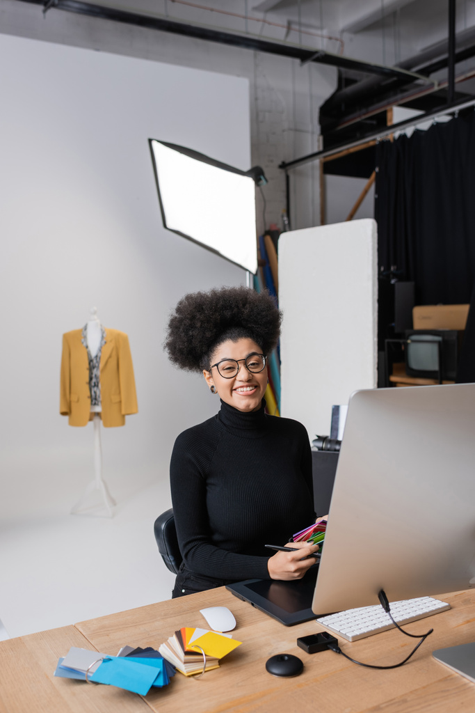 joyeuse retouche afro-américaine en regardant la caméra tout en travaillant avec des échantillons de couleur près de tablette graphique et ordinateur en studio photo - Photo, image