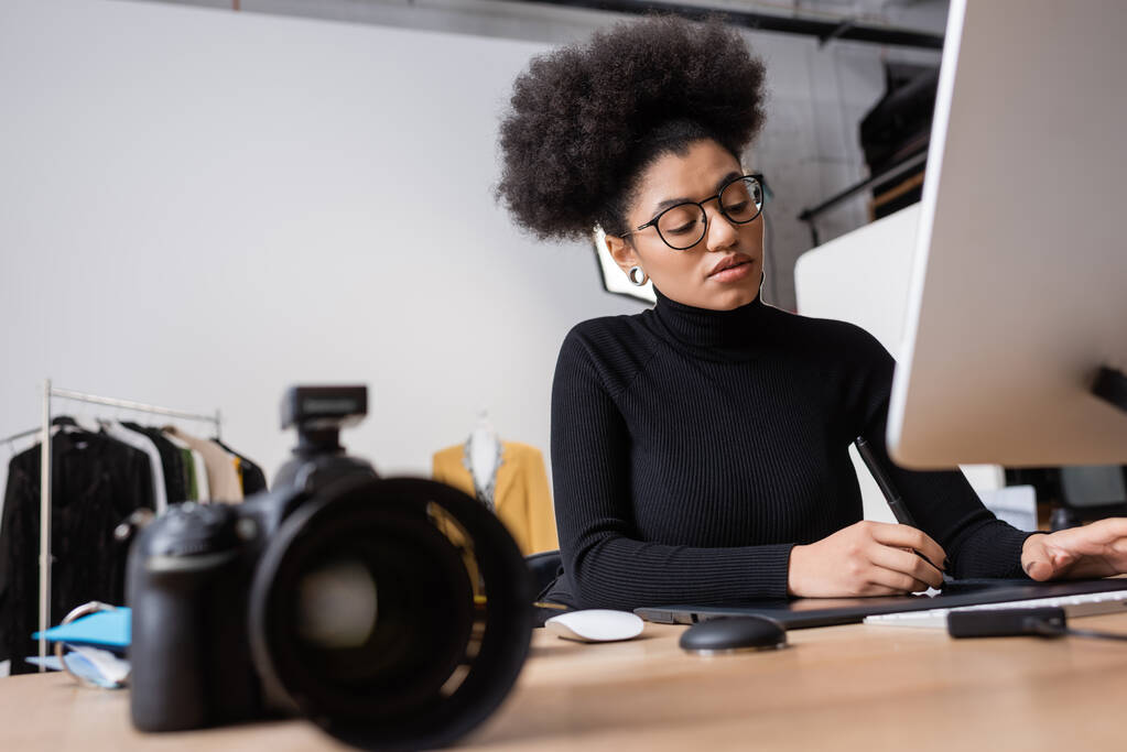 Afro-Amerikan rötuşçusu bilgisayarın yanında grafik tablet kullanıyor ve fotoğraf stüdyosunda bulanık dijital kamera kullanıyor. - Fotoğraf, Görsel