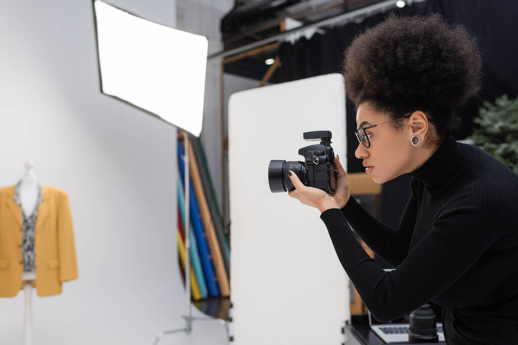 side view of african american photographer with digital camera photographing trendy blazer in photo studio - Photo, Image