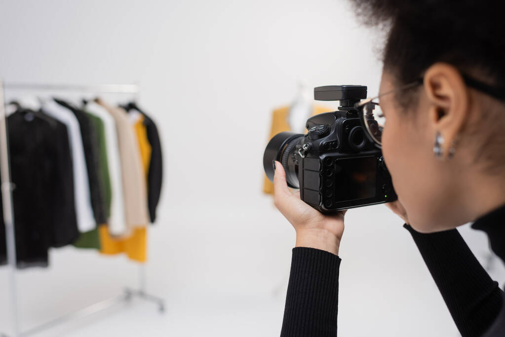 Αφροαμερικανός φωτογράφος με ψηφιακή φωτογραφική μηχανή φωτογραφίζοντας θολή συλλογή ρούχων στο φωτογραφικό στούντιο - Φωτογραφία, εικόνα