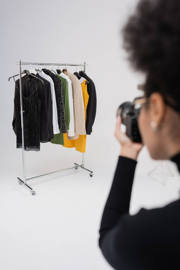 wazig Afrikaans Amerikaans fotograaf neemt foto van stijlvolle kleding op rail rack in fotostudio - Foto, afbeelding