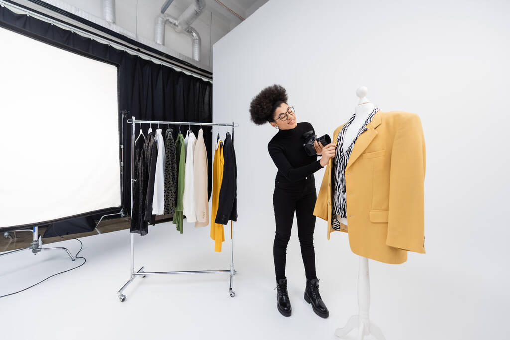 full length of african american content producer with digital camera adjusting trendy blazer on mannequin in photo studio - Photo, Image