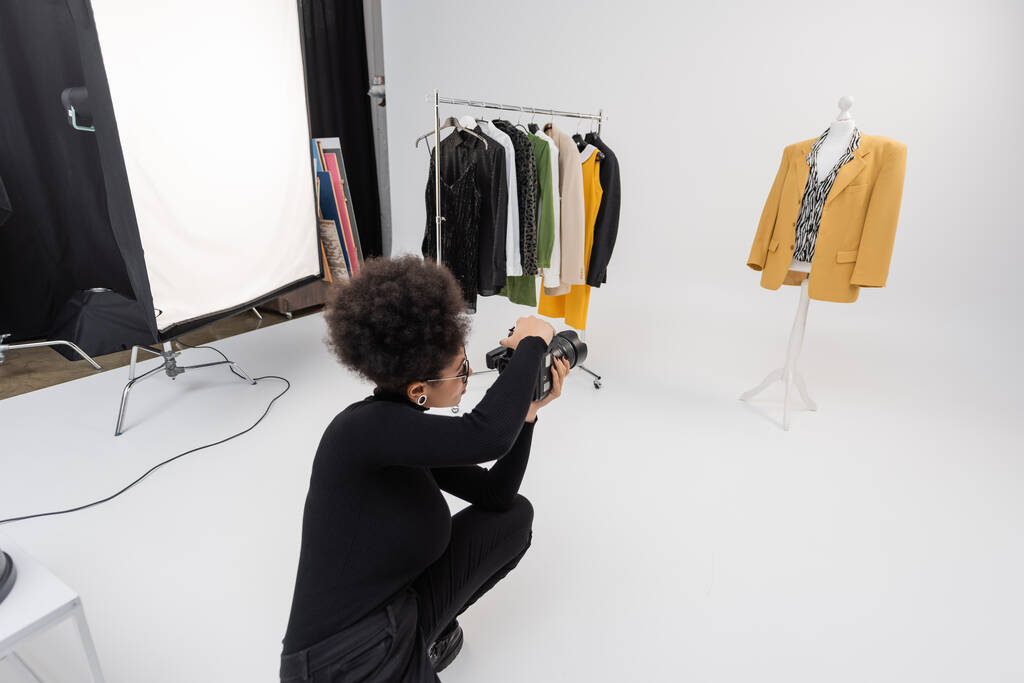 fabricante de conteúdo americano africano com câmera digital perto de roupas da moda no rack ferroviário e manequim no estúdio de fotos - Foto, Imagem