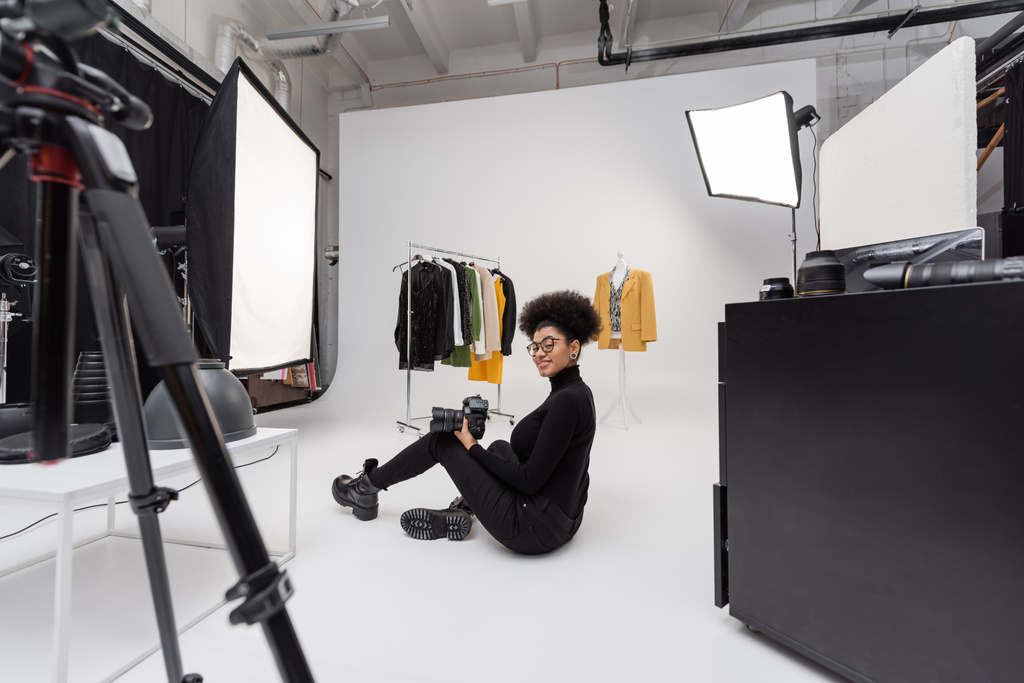 longitud completa de alegre afroamericano fotógrafo mirando a la cámara mientras está sentado cerca de la colección de ropa con estilo en el estudio de fotos - Foto, imagen