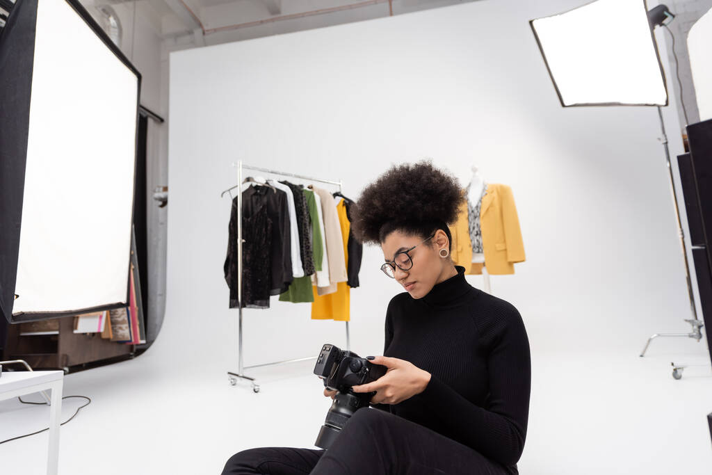 africano americano content maker guardando fotocamera digitale mentre seduto vicino a vestiti alla moda in studio fotografico - Foto, immagini