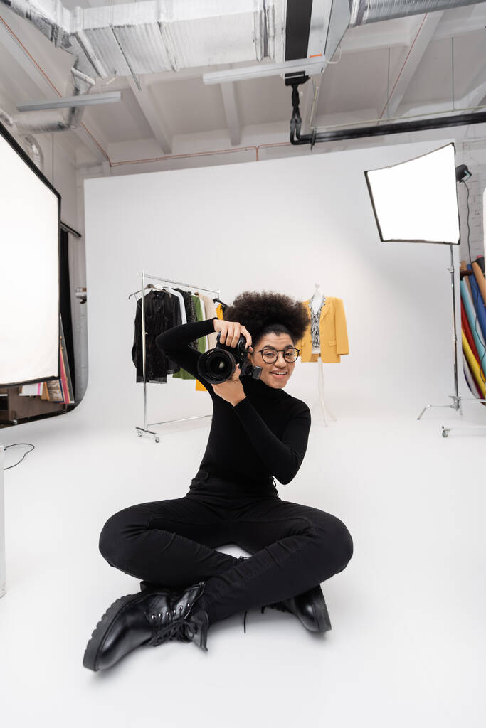 mujer afroamericana feliz tomando la foto en la cámara digital mientras que está sentado en el estudio de la foto cerca de la ropa de moda y focos - Foto, imagen