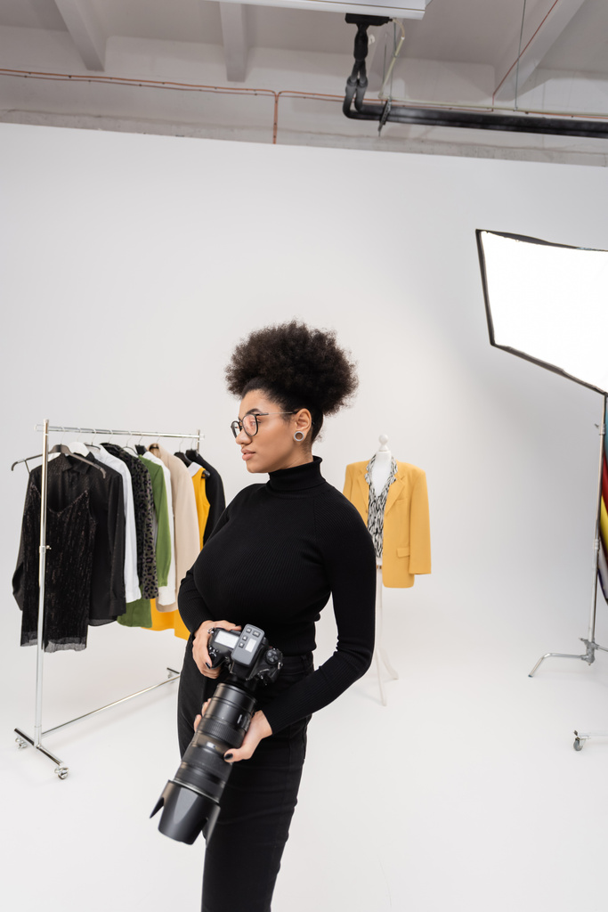 fotógrafo afroamericano en gafas y cuello alto negro de pie con cámara digital cerca de la colección de ropa de moda en el estudio de fotografía - Foto, imagen