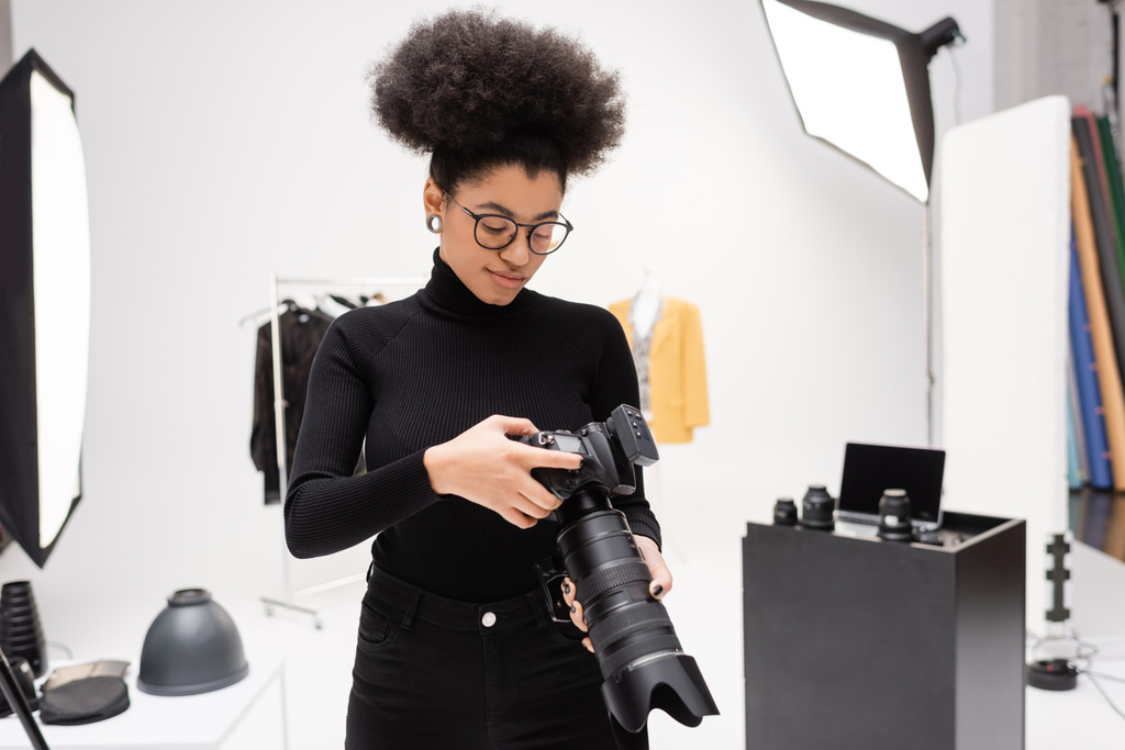 Afrikaans Amerikaans fotograaf in bril en zwarte coltrui op zoek naar professionele digitale camera in fotostudio - Foto, afbeelding